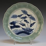A Japanese Arita dish, 18th Century, the flower form celadon rim framing a central panel painted in shades of blue with wind-tossed reeds on a shore with waves and spume clawing the air, pseudo six character mark of Chenghua in underglaze blue to base, diameter approx 26cm.