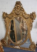 A mid-19th Century giltwood and gesso overmantel mirror surmounted by shell, putti, flower swags and trellis panels above an oval bevelled plate glass and two smaller panels within acanthus leaf borders, height approx 162cm, width approx 132cm (some faults).